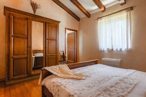 A bedroom with a wooden wardrobe, a bed with a polka dot bedspread, a window with sheer curtains, and a radiator.