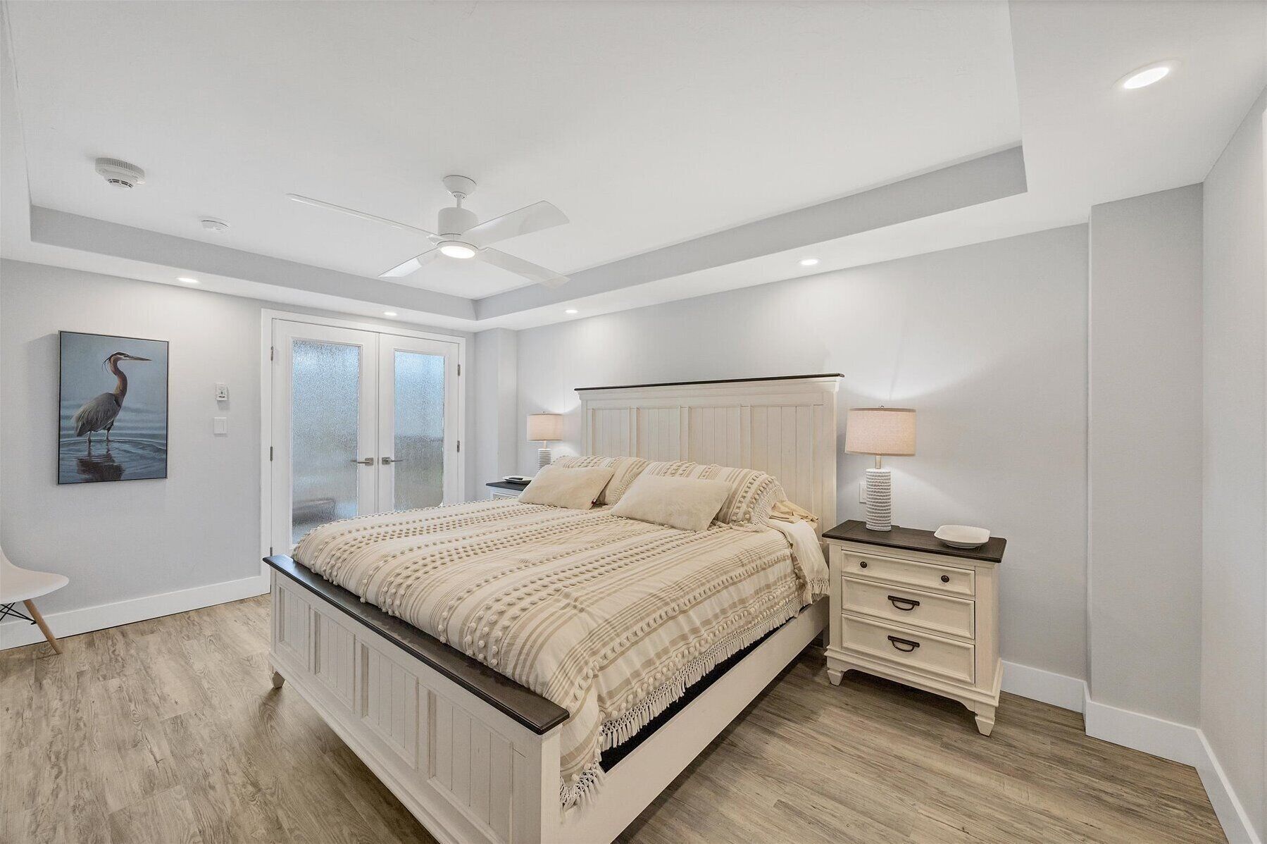 A bedroom with a large bed, a nightstand, a lamp, a ceiling fan, and a door with frosted glass panels.