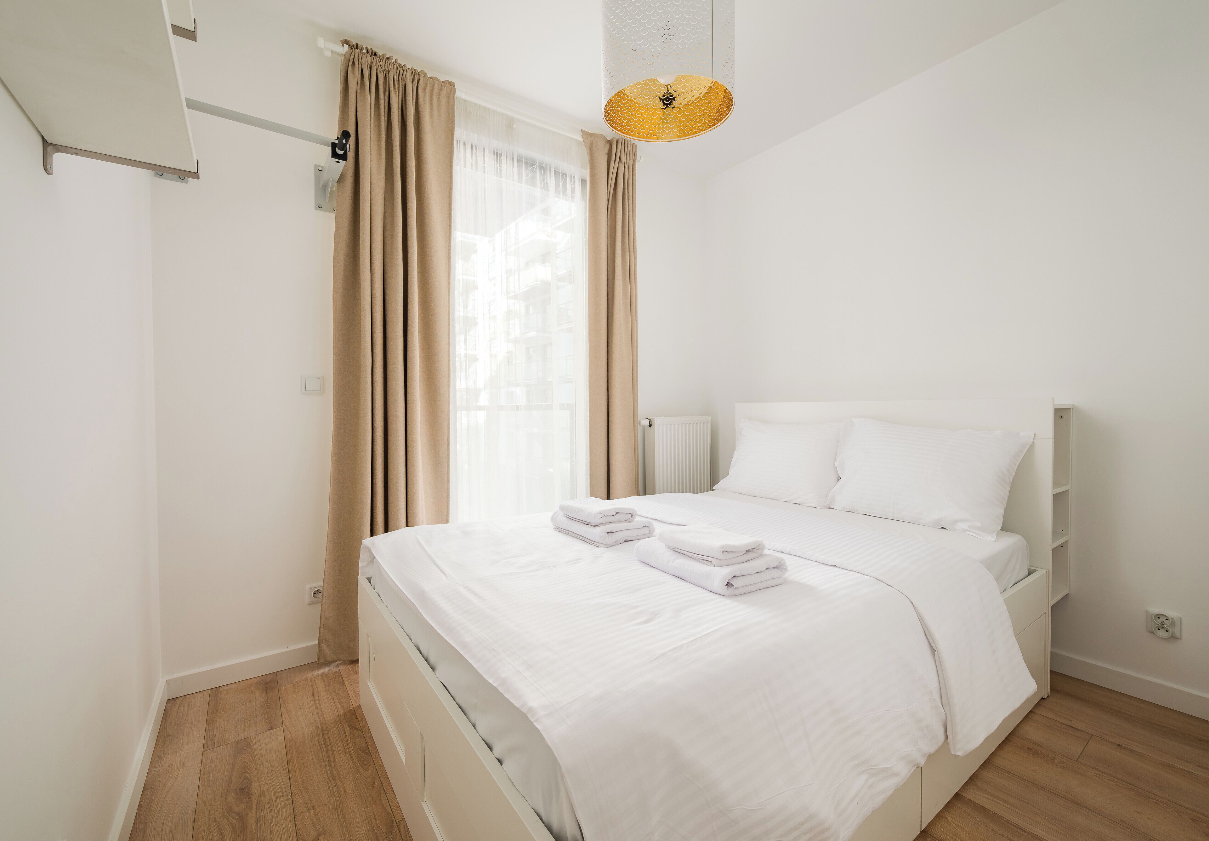 A neatly made bed with white linens and towels, a bedside table, a hanging light fixture, and a window with curtains.