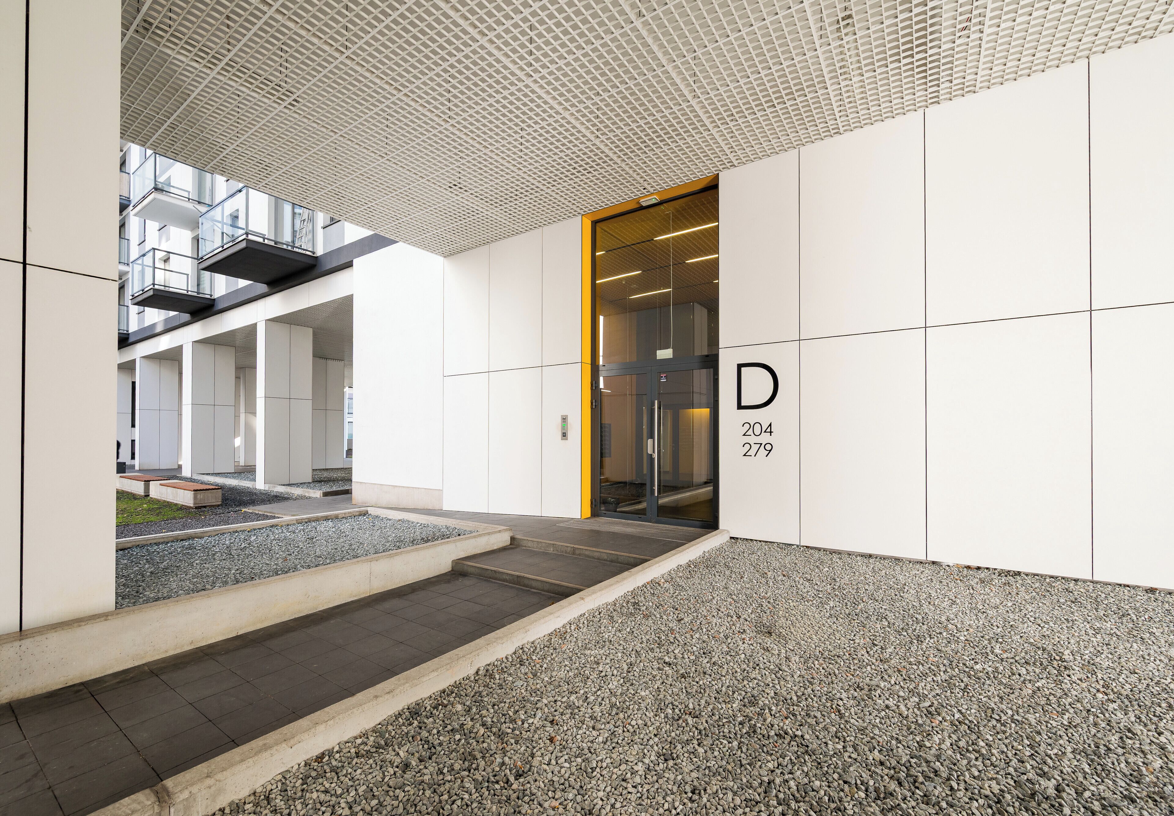 Una entrada moderna a un edificio con puerta de vidrio identificada con 'D 204 279' y un sendero de grava que la conduce.