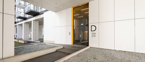 Una entrada moderna a un edificio con puerta de vidrio identificada con 'D 204 279' y un sendero de grava que la conduce.