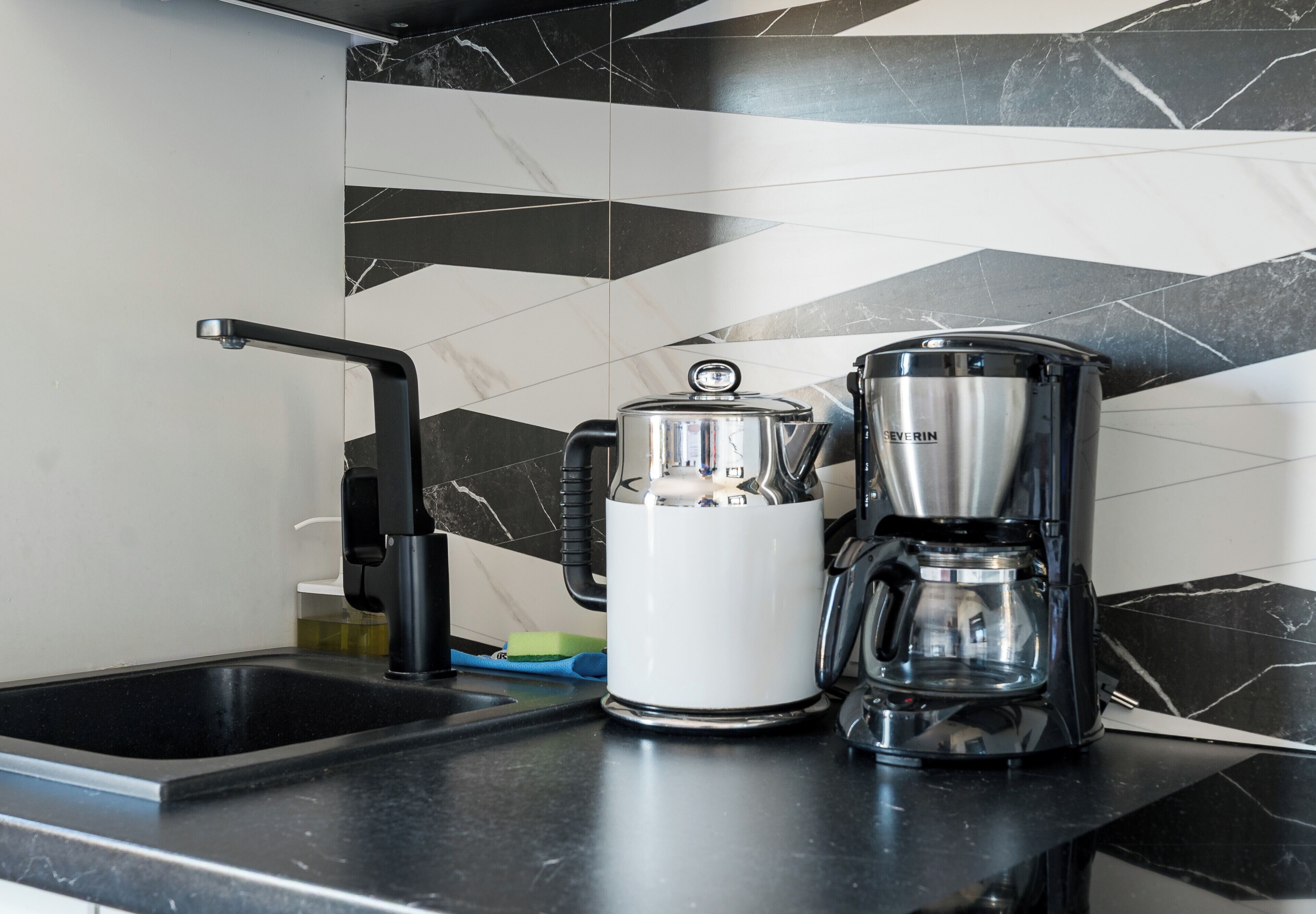Una cocina con un salpicadero blanco y negro de estilo moderno, una tetera de acero inoxidable y una cafetera sobre la encimera.