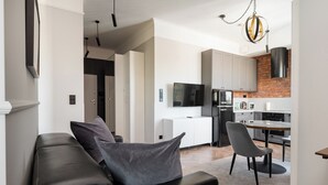A modern living room with a black leather sofa, a dining area with a wooden table, and a kitchen with white cabinets and a brick accent wall.