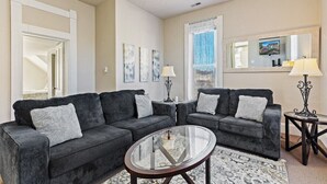 Un salon avec deux canapés gris foncé, une table basse en verre et un tapis à motifs. On y trouve des lampes fixées au mur et un ventilateur de plafond.