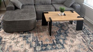 Un salon avec un canapé d’angle gris, une table basse en bois, un tapis à motifs et deux tableaux représentant des fleurs accrochés au mur.