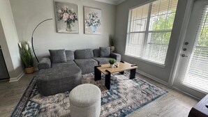 Un salon avec un canapé d’angle gris, une table basse en bois, un tapis à motifs et deux tableaux accrochés au mur.
