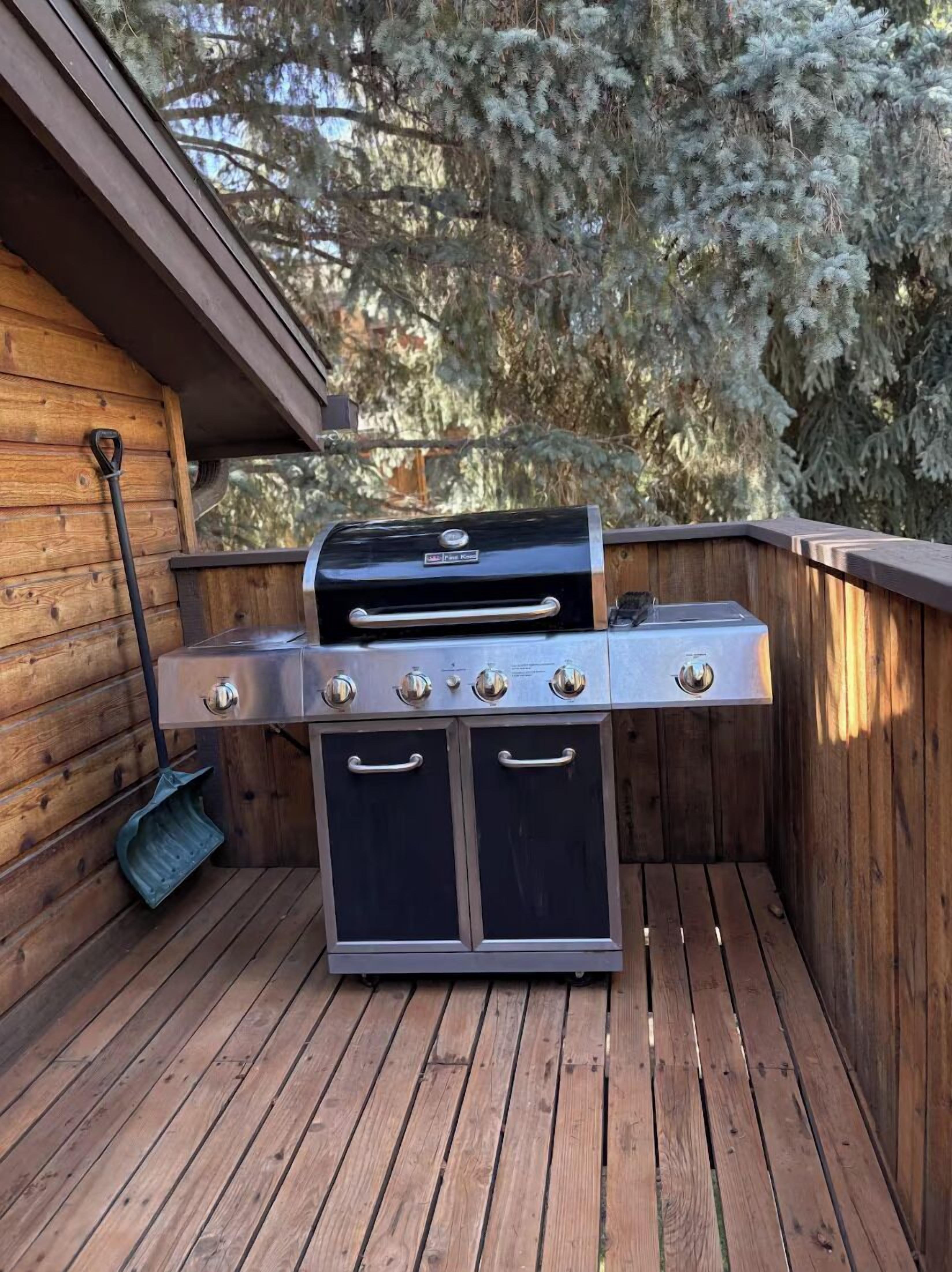 Una griglia barbecue con quattro fuochi, un piano di cottura laterale e un armadietto portaoggetti, posizionata su un pavimento di legno con un muro di legno e alberi sullo sfondo.