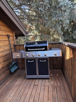 Una griglia barbecue con quattro fuochi, un piano di cottura laterale e un armadietto portaoggetti, posizionata su un pavimento di legno con un muro di legno e alberi sullo sfondo.
