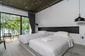 A modern hotel room with two beds, a large window with a view of palm trees, a balcony with a table and chairs, and a black pendant light.