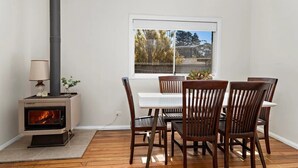 A dining area with a wood-burning stove, wooden chairs, a white table, and a window with a view of trees.