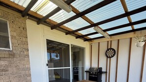 A wooden table and bench set in a covered outdoor area with a brick wall and a grill.