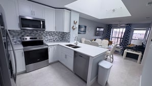 A modern kitchen with white cabinetry, a microwave, stove, and a dishwasher. There is a dining area with a table and chairs, and a small island with a sink. - Picture Perfect: Tilghman Beach & Racquet Club, Steps to the Beach (North Myrtle Beach)