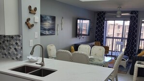 A modern kitchen with a built-in island, a dining area with white chairs, and a large window with a view. - Picture Perfect: Tilghman Beach & Racquet Club, Steps to the Beach (North Myrtle Beach)