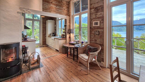 A cozy kitchen with wooden flooring, a fireplace, and large windows offering a scenic view of mountains and water. - Awesome home in Vaksdal with house sea view (Vaksdal)
