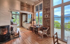 A cozy kitchen with wooden flooring, a fireplace, and large windows offering a scenic view of mountains and water. - Awesome home in Vaksdal with house sea view (Vaksdal)