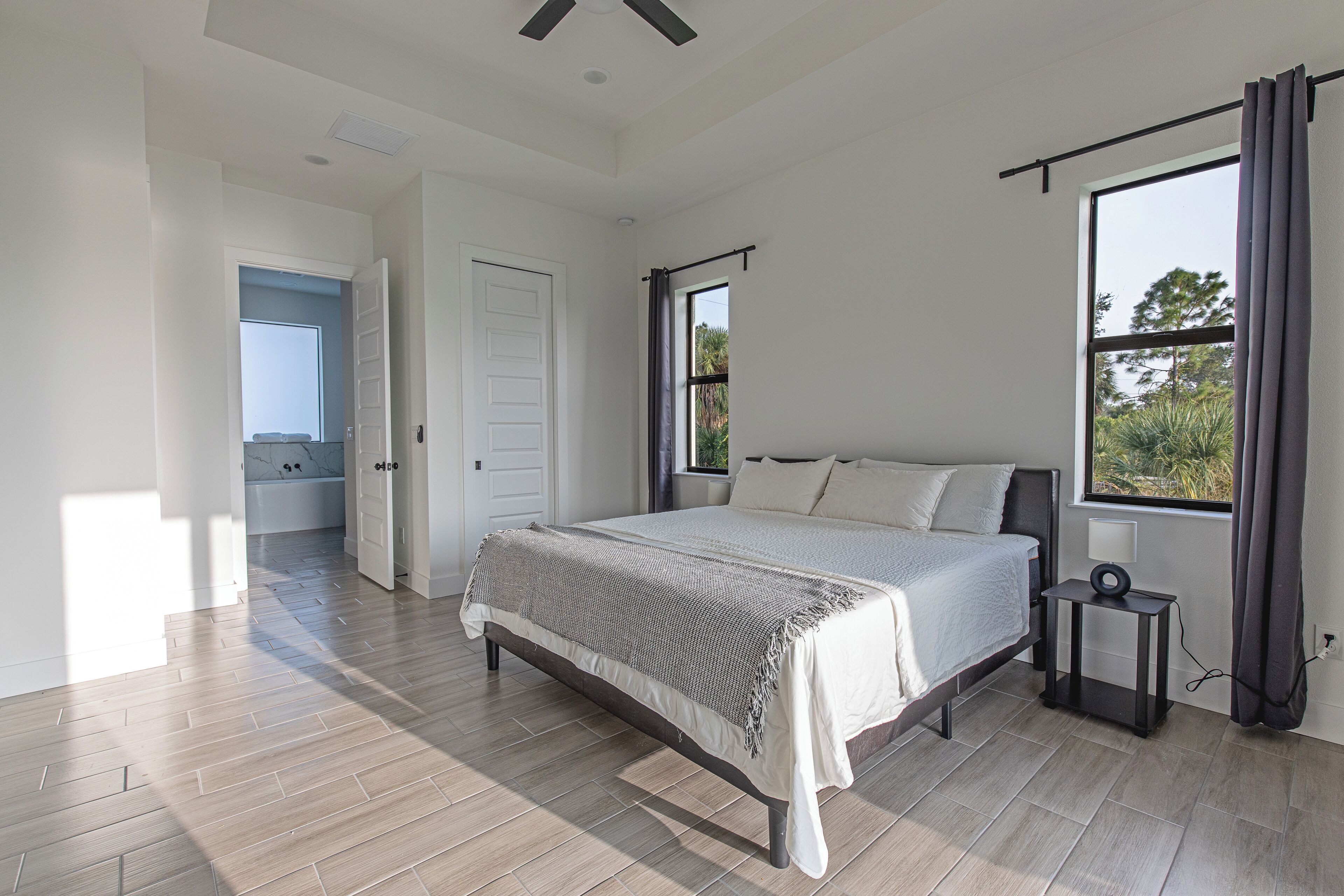 A modern bedroom with a large bed, a small bedside table, a window with curtains, and a view of the outdoors.