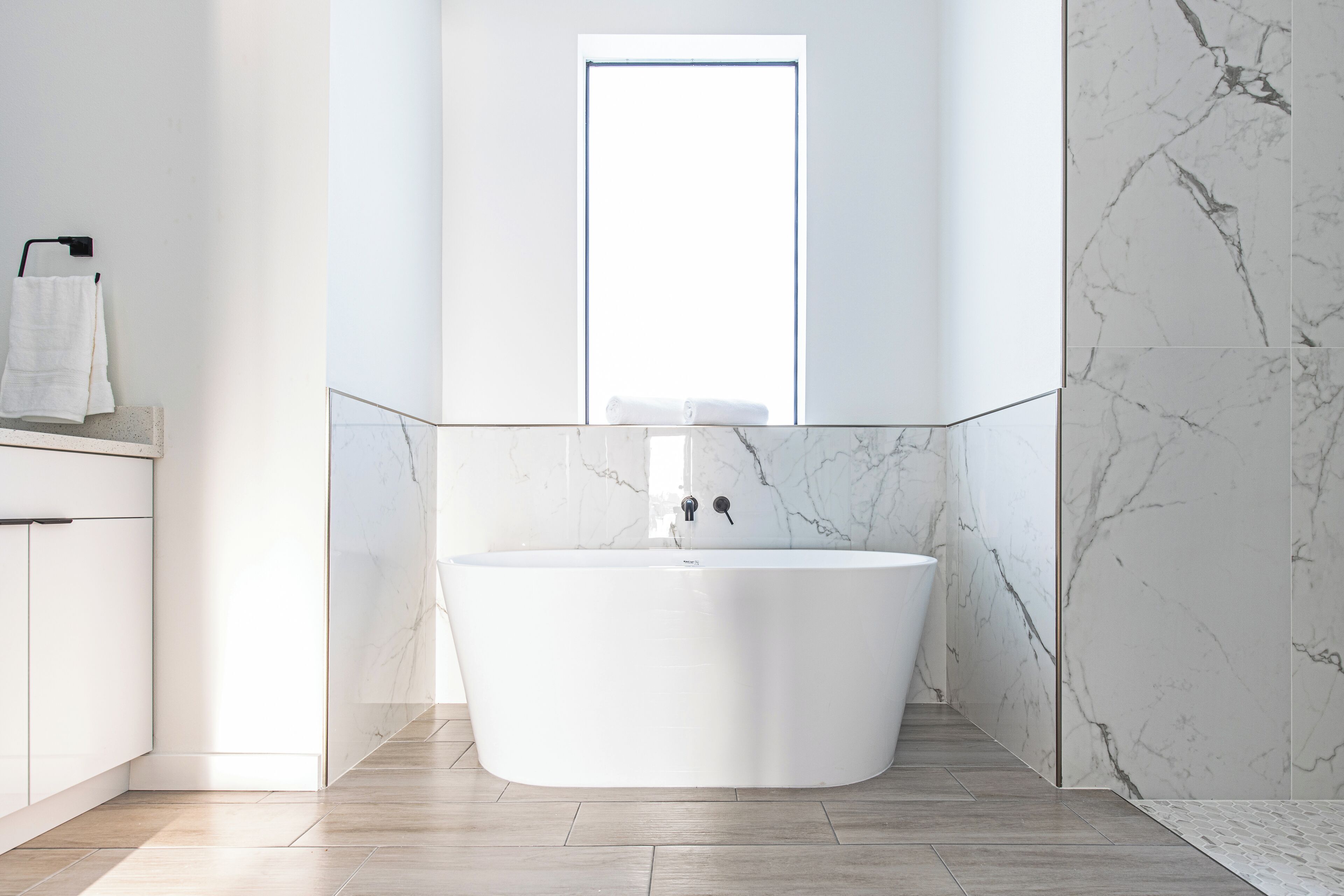 A modern bathroom with a freestanding bathtub, a window, and marble walls.