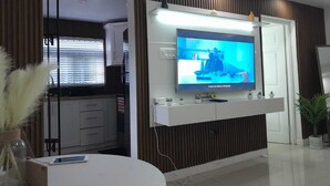 A modern living room with a flat-screen TV mounted on a wooden wall, a dining area with a round table, and a kitchen visible through a glass partition.