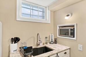 A kitchen with a sink, faucet, and a microwave. There is a window above the sink and a light fixture on the wall.