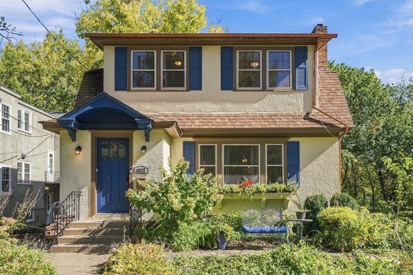A two-story house with a front porch, a blue door, and a small garden with a bench.