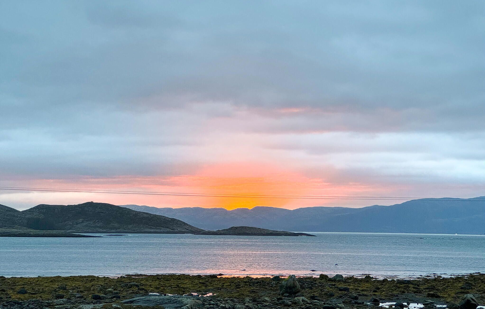 A coastal landscape with a body of water, rocky shoreline, and distant mountains under a cloudy sky.