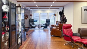 A spacious living room with a wooden floor, leather sofas, a large window with blinds, a mounted ship's wheel, and a mounted animal head.