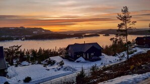 A house with a dark roof, surrounded by snow-covered ground and trees, overlooking a river with a sunset in the background. - Nice home in Sannidal with lake view (Sannidal)