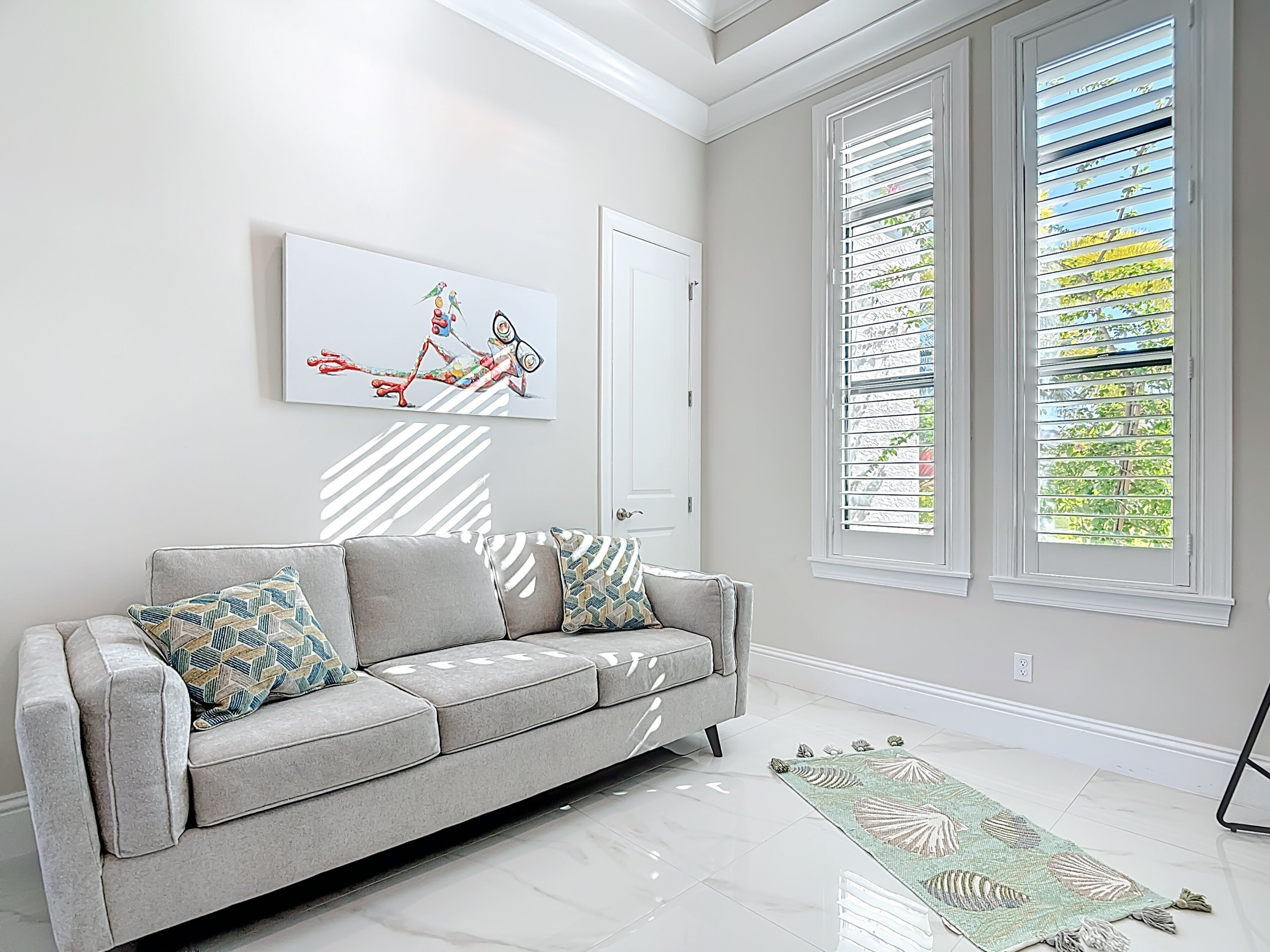 Een moderne woonkamer met een grijze bank, decoratieve kussens, een groot abstract schilderij en uitzicht door horizontale zonwering.