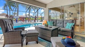 Área ao lado da piscina com mobiliário de vime, uma mesa de jantar e vista para palmeiras e casas.