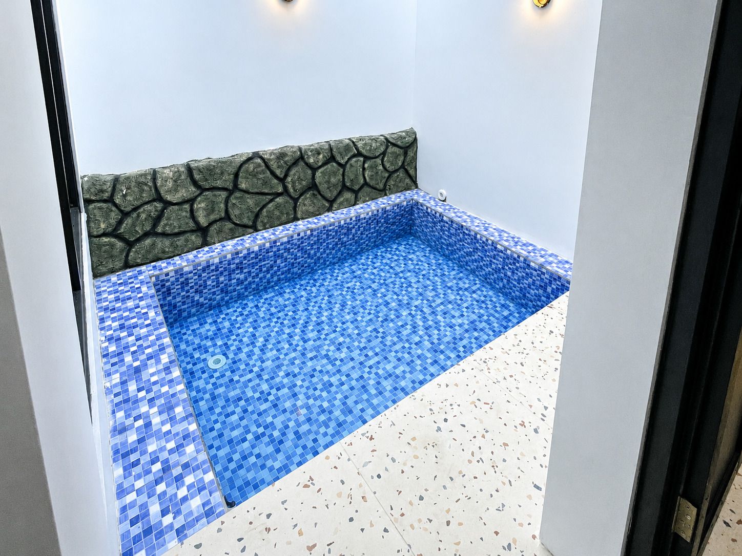A small, square-shaped hot tub with blue mosaic tiles, a stone wall, and a terrazzo floor.