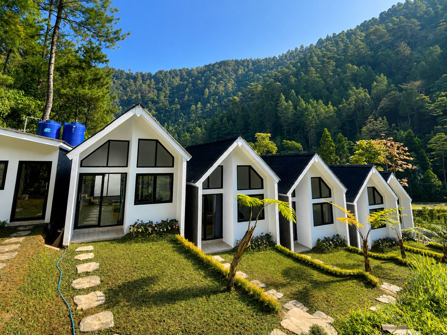 A row of white cabins with black roofs, set against a backdrop of a forested hillside.