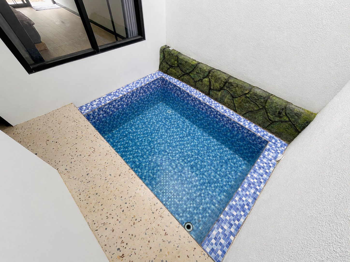 A small, square-shaped pool with a blue mosaic tile border, adjacent to a white wall and a window.