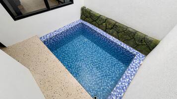 A small, square-shaped pool with a blue mosaic tile border, adjacent to a white wall and a window.