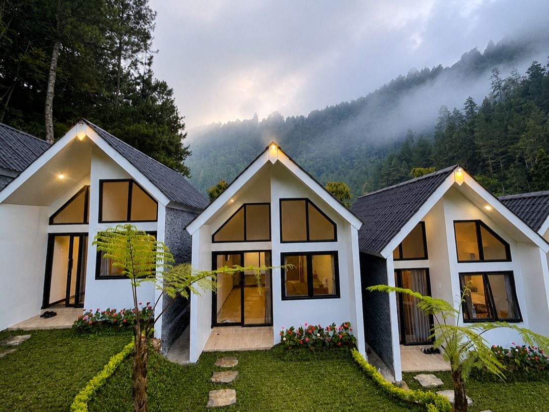 Fileira de casas modernas, brancas, com dois andares, grandes janelas e telhado inclinado, cercada por um jardim bem cuidado e uma montanha arborizada ao fundo.