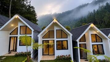 Fileira de casas modernas, brancas, com dois andares, grandes janelas e telhado inclinado, cercada por um jardim bem cuidado e uma montanha arborizada ao fundo.