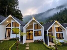 Filas de casas modernas, de color blanco y dos pisos, con amplias ventanas y techo inclinado, rodeadas por un jardín cuidado y un paisaje montañoso cubierto de bosque.