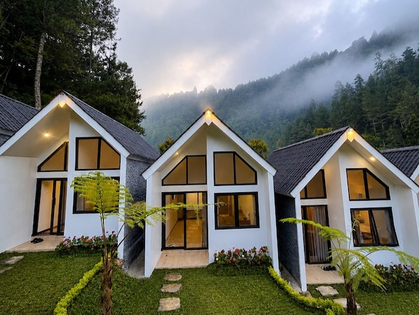 A row of modern, white, two-story houses with large windows and a sloped roof, surrounded by a well-maintained garden and a forested mountain backdrop.