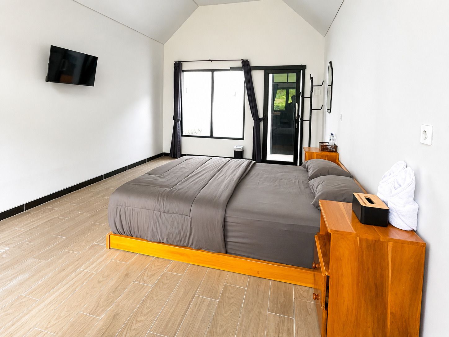 Un dormitorio con una cama, una mesita de noche de madera, un televisor y un ventanal corredero que da a un espacio exterior.