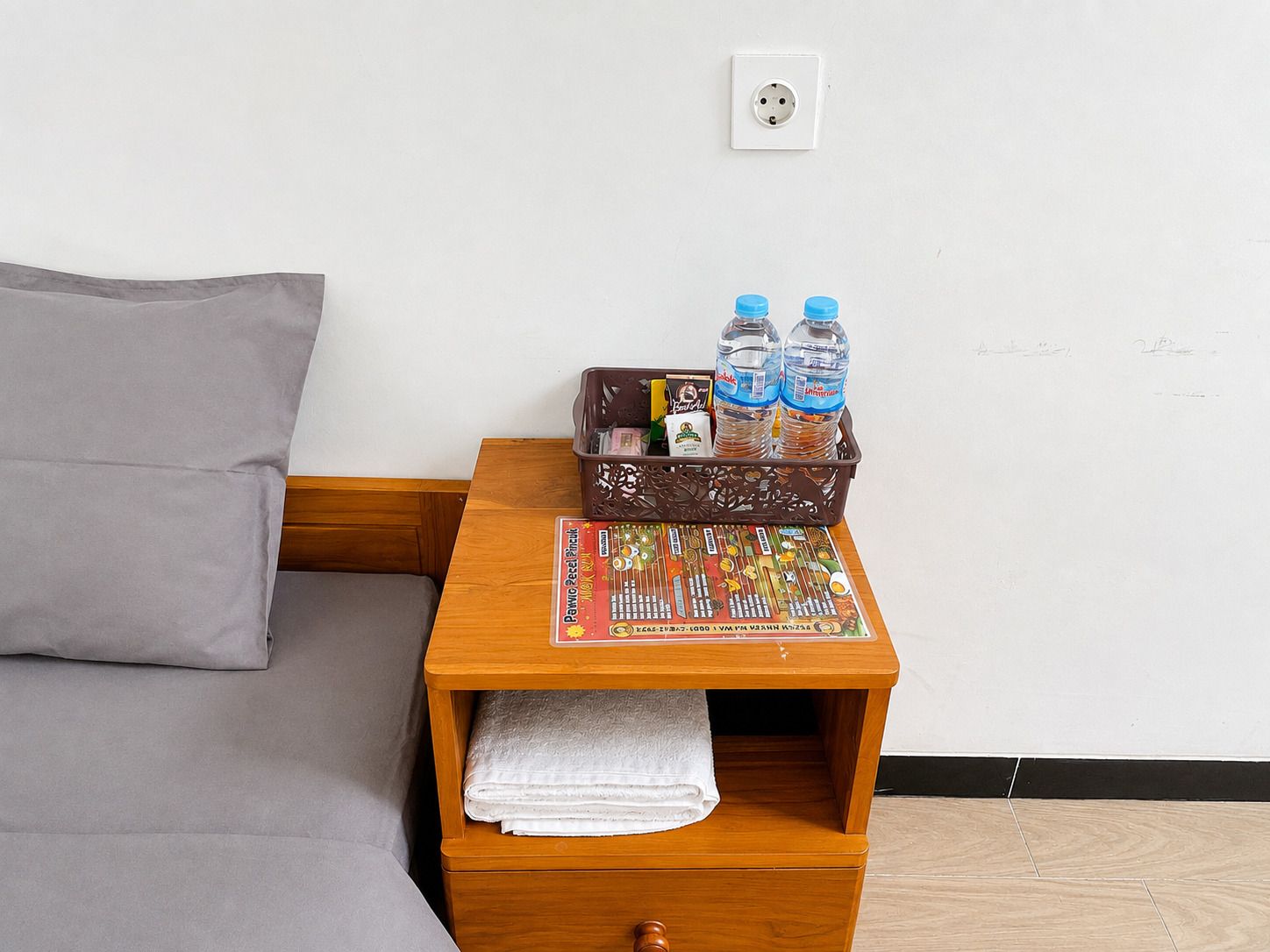 Een net opgemaakt bed met een grijze deken, een houten nachtkastje met een tijdschrift, een fles water en een klein mandje met spullen.