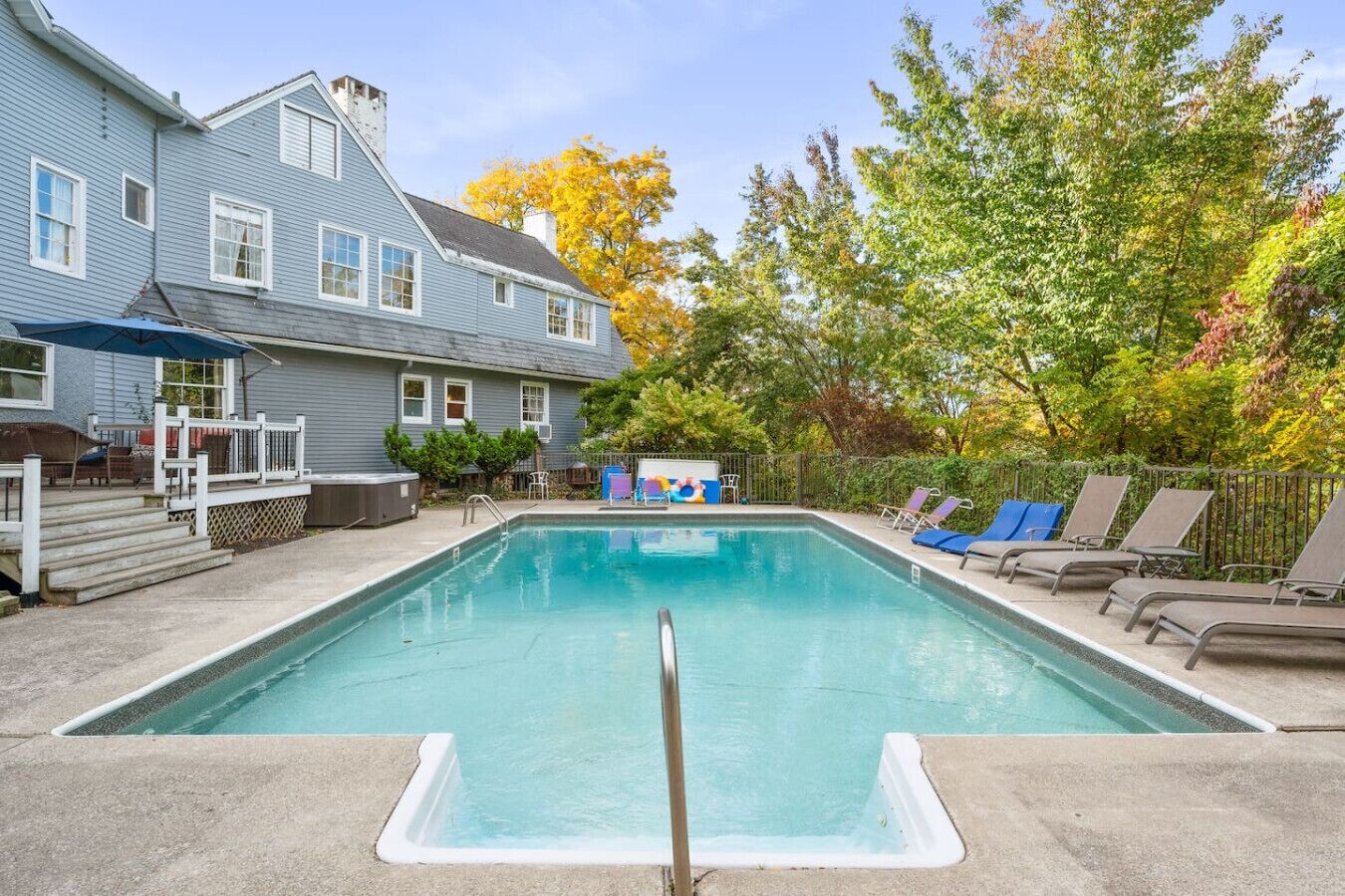 A rectangular swimming pool with a deck area, surrounded by a fence, and a house with a porch in the background.