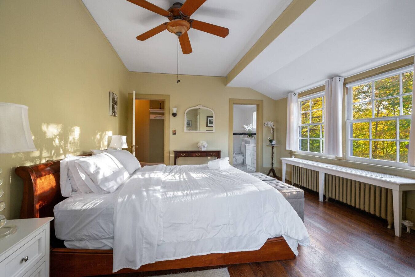 Un dormitorio con cama de madera, ventilador de techo, ventana con vista a árboles, radiador y espejo en la pared.