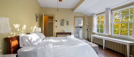 Un dormitorio con cama de madera, ventilador de techo, ventana con vista a árboles, radiador y espejo en la pared.