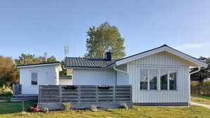 Ein modernes, eingeschossiges Haus mit weißer Fassade, dunklem Dach und Schornstein. Es besitzt eine kleine Veranda mit Treppen und einen Holzzaun.