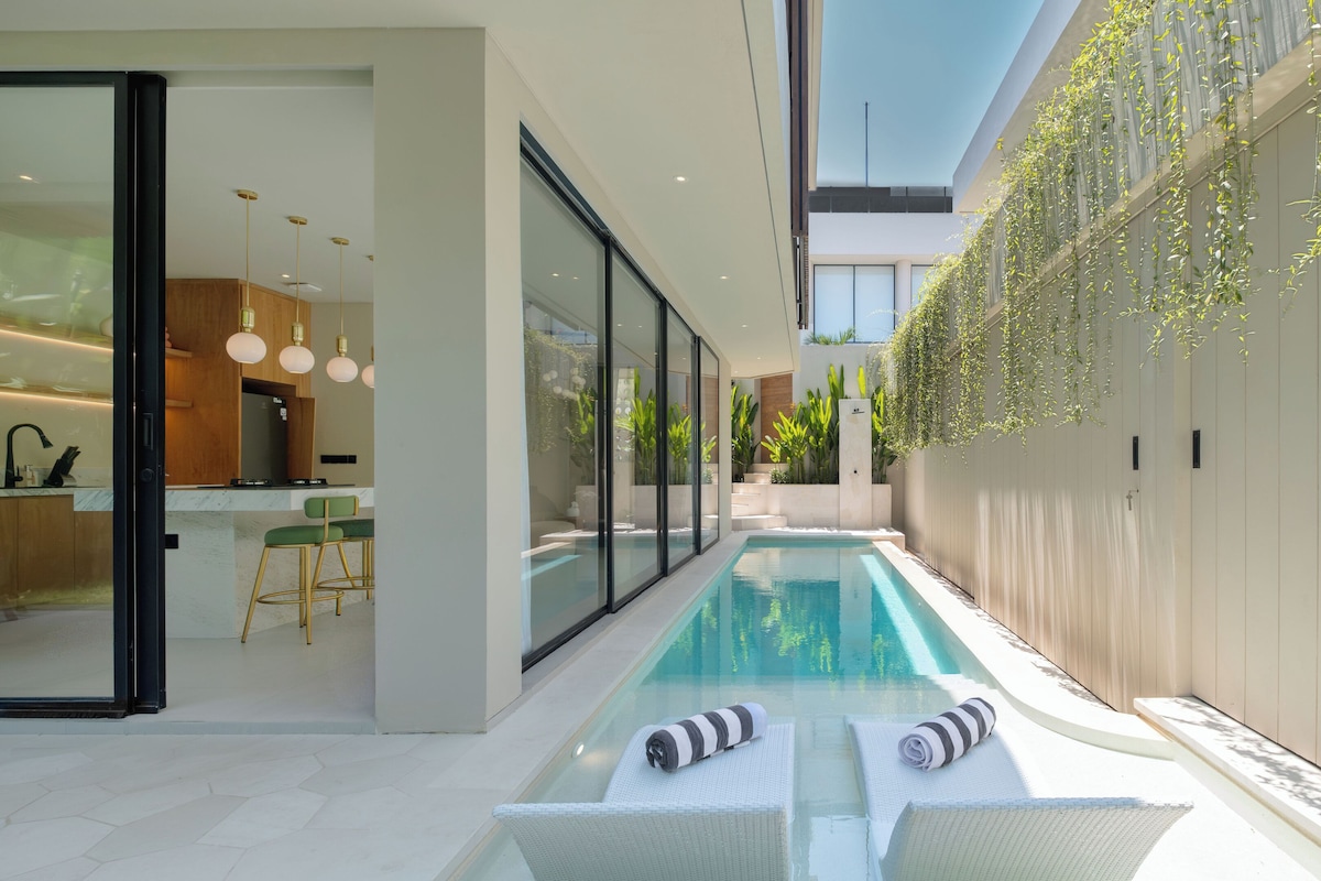 A modern outdoor pool area with glass walls, a kitchen visible through the glass, and a staircase leading to a higher level.