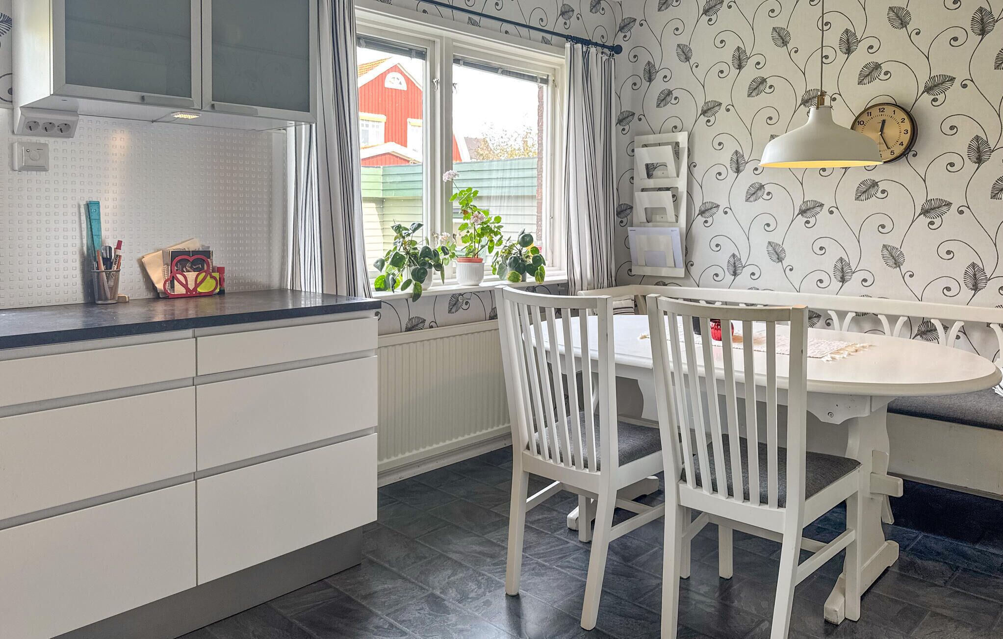 Ett kök med vita skåp, ett fönster med gardiner, ett matbord med stolar och en väggklocka.