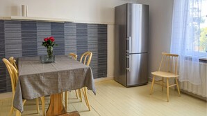 A dining area with a table set for four, a tall refrigerator, and a window with sheer curtains.