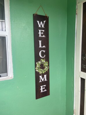 A vertical welcome sign with the letters "WELC ME" and a wreath on a green wall.
