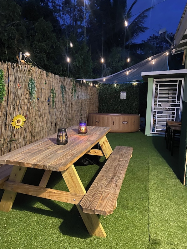 Una mesa de picnic de madera con bancos, rodeada de césped artificial, una valla de bambú y luces colgantes.
