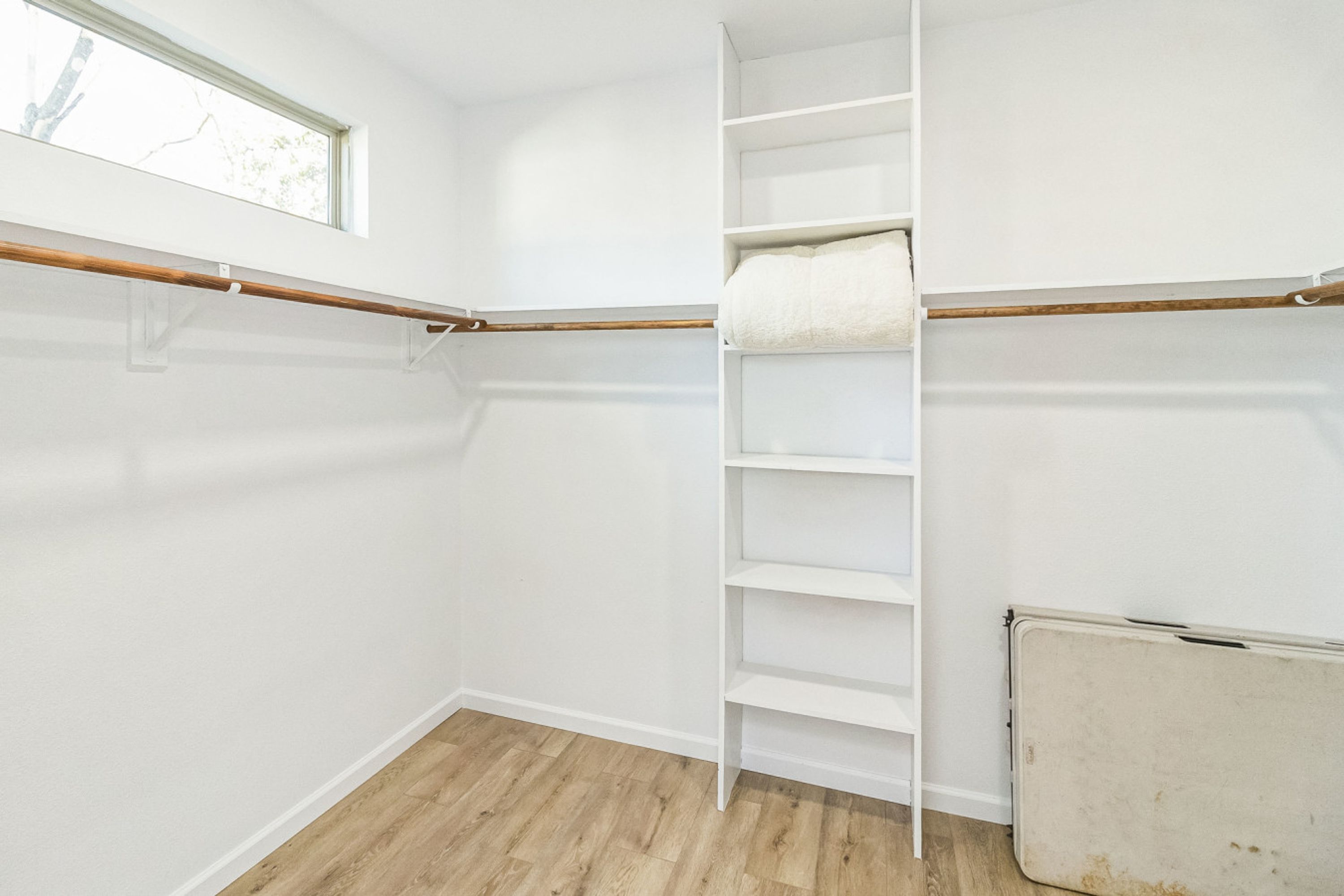 A walk-in closet with wooden flooring, white walls, and a window.