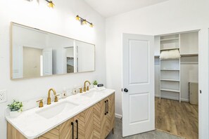 A bathroom with a white vanity, gold faucets, a large mirror, and a walk-in closet with shelves. - Stylish 4BR Home Family-friendly Near Downtown (San Antonio)